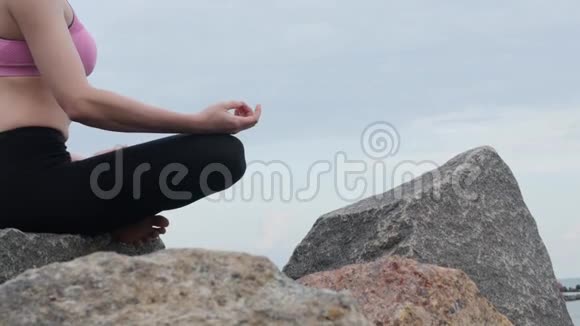 女孩练习瑜伽健身运动靠近海边坐在石头上的莲花姿势视频的预览图