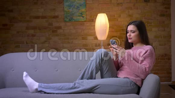 一位年轻漂亮的黑发白种人女性她用电话和微笑悠闲地坐在沙发上视频的预览图