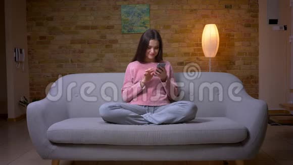 一位年轻漂亮的褐发白种人女性在舒适的沙发上用电话微笑特写镜头视频的预览图