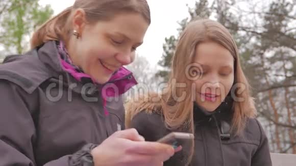 两个快乐的女孩母亲和女儿坐在公园里和电话交谈4KUHD视频的预览图