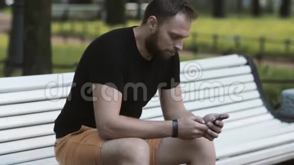在公园里坐在长椅上使用智能手机的一位身材矮小的年轻人视频的预览图