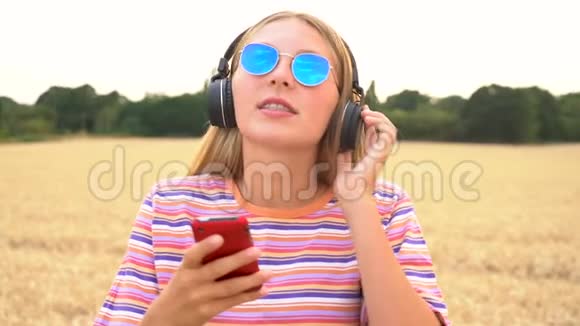 年轻女子戴着蓝色太阳镜在野外听无线耳机音乐视频的预览图
