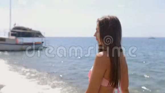 迷人的年轻女子长着一头直发穿着粉红色比基尼沿着海滩的海岸散步视频的预览图