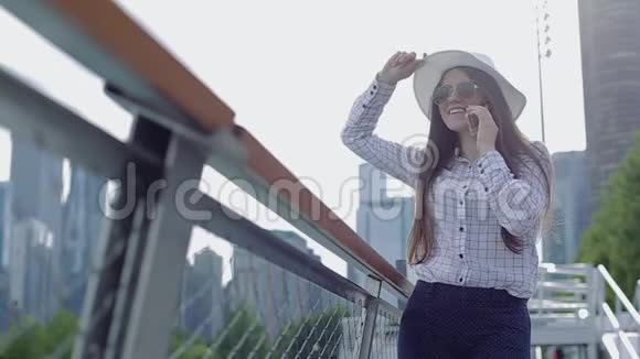 一位戴着帽子和太阳镜的年轻女子站在阳光下的长廊上打电话视频的预览图