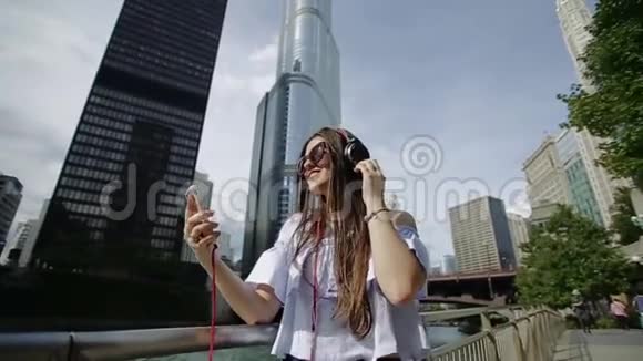 年轻美丽的女人听着耳机在河堤上跳舞有摩天大楼和运河视频的预览图