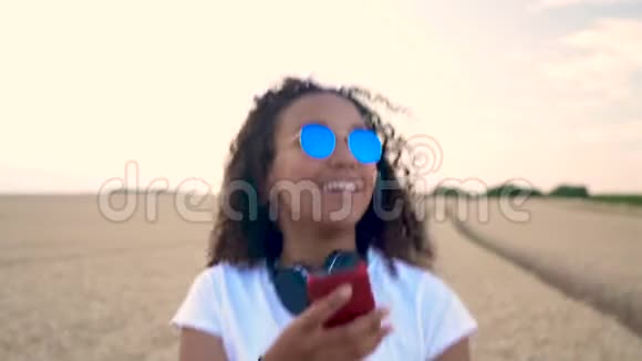 戴着蓝色太阳镜的年轻女子走在田野里用无线耳机听音乐视频的预览图