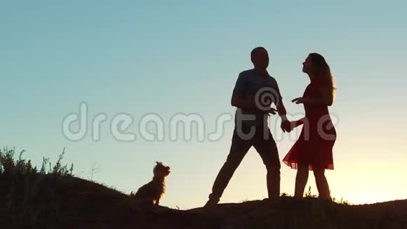 一对幸福的年轻夫妇的剪影和日落时在外面慢舞的狗慢动作视频男人和女孩视频的预览图