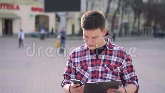 年轻的帅哥在阳光明媚的街道上用笔记本和电话说话视频的预览图