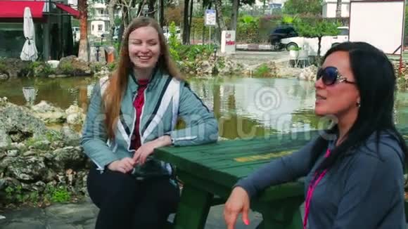 两个年轻开朗的女人坐在长凳上在公园的一张桌子旁说话视频的预览图