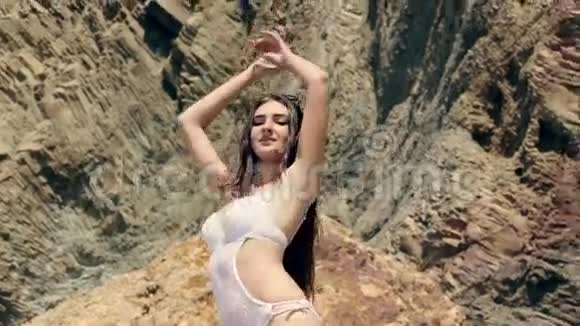 美丽的年轻女子一头乌黑的长发身穿白色泳衣站在沙滩上的岩石上跳舞视频的预览图