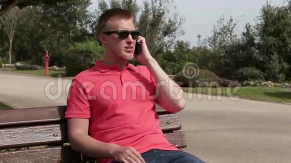 夏天一个快乐的年轻人坐在公园的长凳上用智能手机聊天视频的预览图