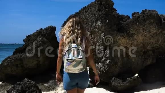 年轻的女孩一头漂亮的头发穿着工作服背着背包在美丽的白色海滩上带着岩石视频的预览图
