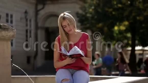 在喷泉附近穿着玫瑰T恤和蓝色牛仔裤看书的漂亮学生视频的预览图