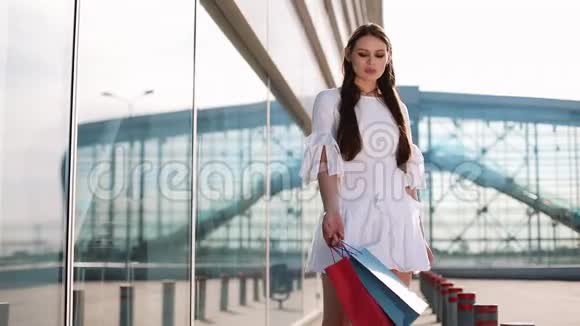 在现代玻璃建筑前穿着白色连衣裙的漂亮时尚模特摆着购物袋视频的预览图