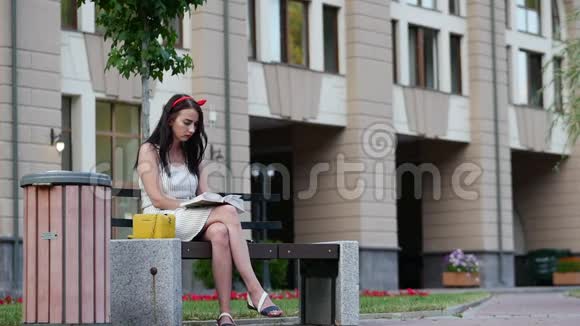 可爱的女孩头上有一个红色的边缘坐在公园的长凳上读一本书视频的预览图