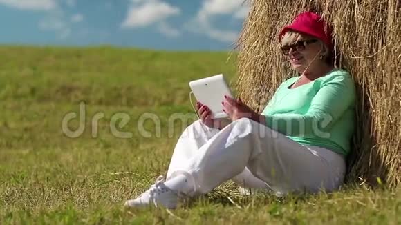 金发女人坐在干草堆附近的绿草上通过平板电脑交流视频的预览图