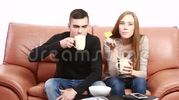 年轻的异性恋夫妇在沙发上拥抱在家里看电视喝茶视频的预览图