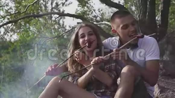 快乐的青少年夫妇在徒步旅行时在森林里的烟熏篝火上吃香肠视频的预览图