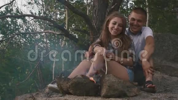 快乐少年夫妇在森林里烟雾弥漫的篝火上烤香肠年轻人高兴地吻他的女朋友视频的预览图