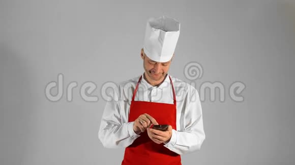 英俊的成人厨师在智能手机上打字然后他看着相机在灰色背景上微笑视频的预览图