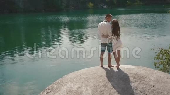 美丽的被爱情侣在湖岸的户外亲吻站在巨大的岩石上视频的预览图