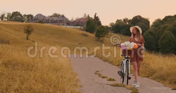 年轻的金发女郎戴着帽子穿着连衣裙带着自行车和花篮走着视频的预览图