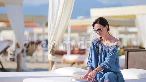 年轻女子在热带海滩度假期间看书时尚女郎坐在欧洲海滩的白色日光浴床上看书视频的预览图