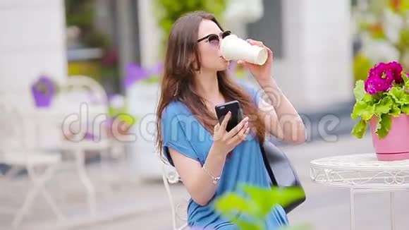 美女坐在户外咖啡馆喝咖啡和使用智能手机的肖像年轻女孩在看视频的预览图