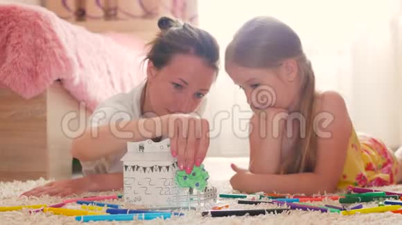 年轻的母亲和她美丽的女儿画一个纸房子躺在家里的地板上生活方式创造力视频的预览图