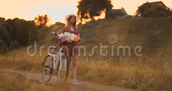 年轻的金发女郎戴着帽子穿着连衣裙带着自行车和花篮走着视频的预览图