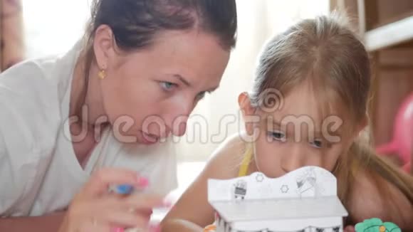 年轻的母亲和她美丽的女儿画一个纸房子躺在家里的地板上生活方式创造力视频的预览图