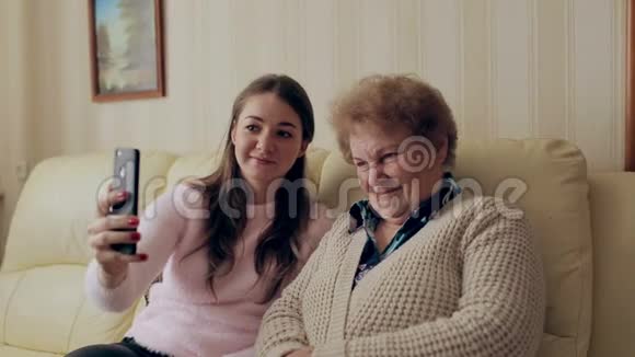 奶奶和年轻女孩在家里坐在沙发或沙发上用手机或智能手机拍照快乐的女士们视频的预览图