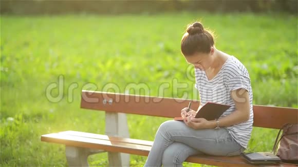 美丽的年轻女子正在公园的户外写日记学生在公园的长凳上学习学生女孩视频的预览图