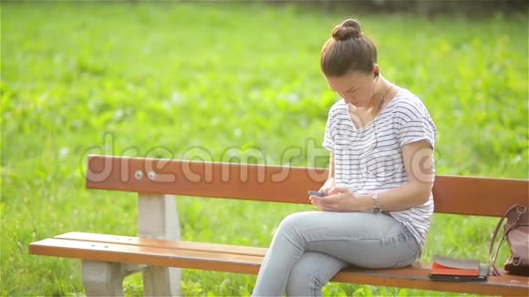 在公园里一个带着耳机听着智能手机音乐的女人一个使用智能手机的漂亮女人视频的预览图