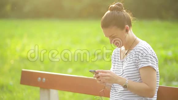 在公园里一个带着耳机听着智能手机音乐的女人一个使用智能手机的漂亮女人视频的预览图