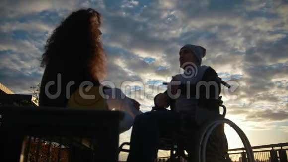 有残疾男子坐轮椅的年轻女子在日落时分在户外聊天视频的预览图