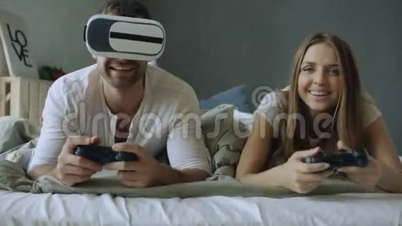 躺在床上的年轻夫妇用控制器和VR耳机玩电子游戏视频的预览图
