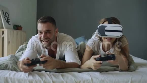 躺在床上的年轻夫妇用控制器和VR耳机玩电子游戏视频的预览图