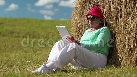金发女人坐在干草堆附近的绿草上使用平板电脑视频的预览图