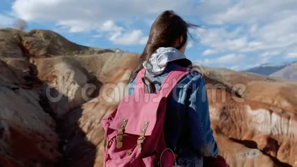 一个穿着牛仔裤夹克和粉红色背包的女孩在刮风的天气特写4k欣赏群山视频的预览图