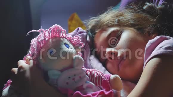 可爱的小女孩睡在沙发床上拥抱她的娃娃晚上睡在床上的黑发小女孩视频的预览图