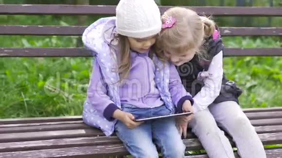 两个拿着平板电脑的小女孩坐在花园的长凳上视频的预览图