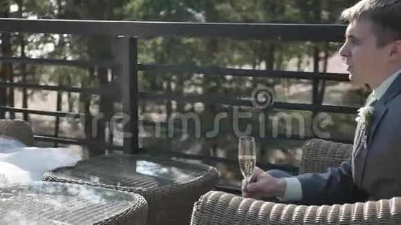 美丽的年轻新郎拿着一杯香槟坐在夏日咖啡馆的阳台上视频的预览图