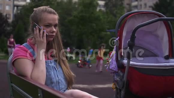 年轻的母亲坐在操场上的长凳上和电话交谈婴儿车夏季视频的预览图