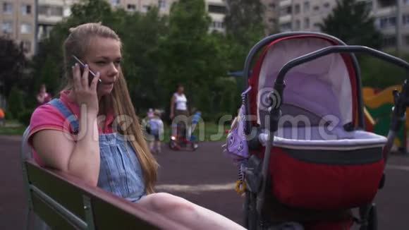 年轻的母亲坐在操场上的长凳上和电话交谈红色婴儿车视频的预览图