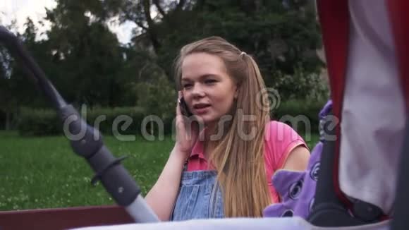 年轻的母亲在长凳上和电话交谈婴儿车夏日微笑视频的预览图