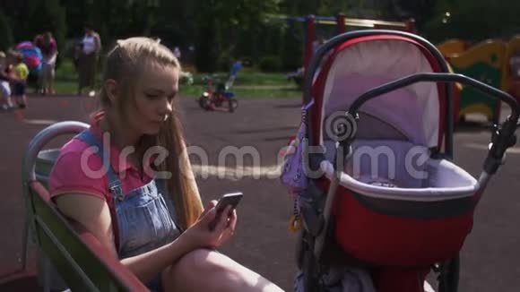 年轻的母亲坐在操场上的长凳上敲着智能手机婴儿车视频的预览图