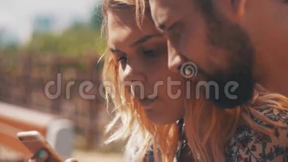 年轻的恋爱情侣坐在长椅上使用智能手机夏日阳光明媚视频的预览图