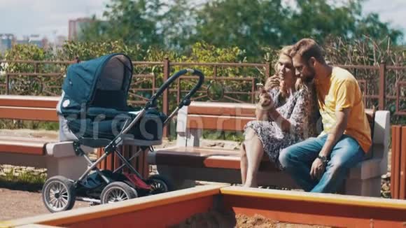 年轻的父母带着婴儿车坐在长凳上使用智能手机家庭游乐场视频的预览图