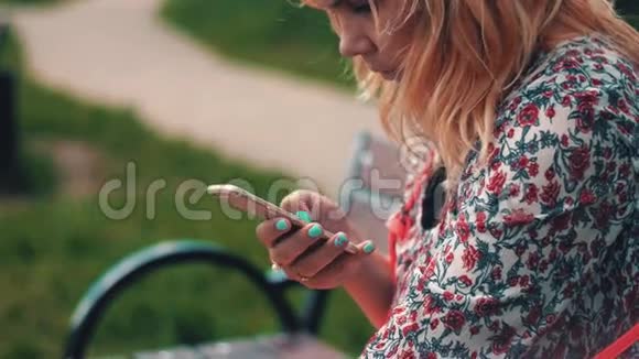 年轻女子坐在长凳上在公园里使用智能手机夏日微笑视频的预览图